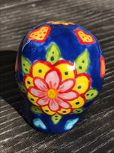 Load image into Gallery viewer, Inspired by nature, this ceramic skull statuette is full of vibrant color and intricate details. The primary color is royal blue with multi-color floral motifs that are painted across the skull, along with hearts and leaves and lively colorful accents around the entire skull.