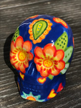 Load image into Gallery viewer, Inspired by nature, this ceramic skull statuette is full of vibrant color and intricate details. The primary color is royal blue with multi-color floral motifs that are painted across the skull, along with hearts and leaves and lively colorful accents around the entire skull.