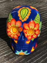 Load image into Gallery viewer, Inspired by nature, this ceramic skull statuette is full of vibrant color and intricate details. The primary color is royal blue with multi-color floral motifs that are painted across the skull, along with hearts and leaves and lively colorful accents around the entire skull.