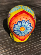 Load image into Gallery viewer, Inspired by nature, this ceramic skull statuette is full of vibrant color and intricate details. The primary color is yellow with Multi-color floral motifs that are painted across the skull, along with hearts and leaves and lively colorful accents around the entire skull.