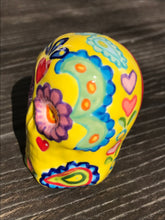 Load image into Gallery viewer, Inspired by nature, this ceramic skull statuette is full of vibrant color and intricate details. The primary color is yellow with Multi-color floral motifs that are painted across the skull, along with hearts and leaves and lively colorful accents around the entire skull.