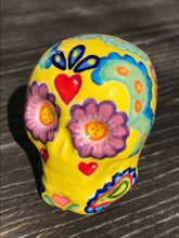Load image into Gallery viewer, Inspired by nature, this ceramic skull statuette is full of vibrant color and intricate details. The primary color is yellow with Multi-color floral motifs that are painted across the skull, along with hearts and leaves and lively colorful accents around the entire skull.