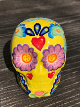 Load image into Gallery viewer, Inspired by nature, this ceramic skull statuette is full of vibrant color and intricate details. The primary color is yellow with Multi-color floral motifs that are painted across the skull, along with hearts and leaves and lively colorful accents around the entire skull.