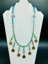 Load image into Gallery viewer, This necklace features vivid green and blue glass beading and a yin yang brass octagon charm. Each side of the charm is distinct, creating a two-in-one look. The look is further enhanced with the addition of rectangular rainbow hematite beads for added visual complexity. Double-hole flat matte gold beads keep the collar shape and add to the necklace's elegant draping when worn.  Measurements:    Necklace is 23” in length