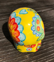 Load image into Gallery viewer, Inspired by nature, this ceramic skull statuette is full of vibrant color and intricate details. The primary color is yellow with Multi-color floral motifs that are painted across the skull, along with hearts and leaves and lively colorful accents around the entire skull.