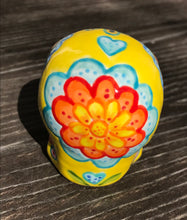 Load image into Gallery viewer, Inspired by nature, this ceramic skull statuette is full of vibrant color and intricate details. The primary color is yellow with Multi-color floral motifs that are painted across the skull, along with hearts and leaves and lively colorful accents around the entire skull.