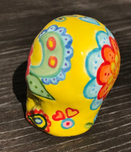 Load image into Gallery viewer, Inspired by nature, this ceramic skull statuette is full of vibrant color and intricate details. The primary color is yellow with Multi-color floral motifs that are painted across the skull, along with hearts and leaves and lively colorful accents around the entire skull. Measurements: H 2 3/4” x W 2” x D 2”