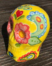 Load image into Gallery viewer, Inspired by nature, this ceramic skull statuette is full of vibrant color and intricate details. The primary color is yellow with Multi-color floral motifs that are painted across the skull, along with hearts and leaves and lively colorful accents around the entire skull. Measurements: H 2 3/4” x W 2” x D 2”