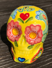 Load image into Gallery viewer, Inspired by nature, this ceramic skull statuette is full of vibrant color and intricate details. The primary color is yellow with Multi-color floral motifs that are painted across the skull, along with hearts and leaves and lively colorful accents around the entire skull. Measurements: H 2 3/4” x W 2” x D 2”