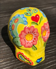 Load image into Gallery viewer, Inspired by nature, this ceramic skull statuette is full of vibrant color and intricate details. The primary color is yellow with Multi-color floral motifs that are painted across the skull, along with hearts and leaves and lively colorful accents around the entire skull. Measurements: H 2 3/4” x W 2” x D 2”