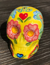 Load image into Gallery viewer, Inspired by nature, this ceramic skull statuette is full of vibrant color and intricate details. The primary color is yellow with Multi-color floral motifs that are painted across the skull, along with hearts and leaves and lively colorful accents around the entire skull.