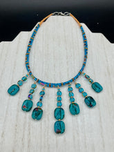 Load image into Gallery viewer, This necklace features vibrant teal quartzite and glass beads. The quartzite beads have a captivating swirl pattern, combining dark red and dark blue hues. It has an ethereal feel thanks to the various shades of blues, golds, and greens used to create a two-strand beaded design. For a dramatic finish, seven strands are added, completed with rectangular rainbow hematite beads for texture and depth. Both strands are held together with double-hole flat matte gold beads to keep a collar shape.