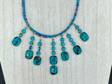 Load image into Gallery viewer, This necklace features vibrant teal quartzite and glass beads. The quartzite beads have a captivating swirl pattern, combining dark red and dark blue hues. It has an ethereal feel thanks to the various shades of blues, golds, and greens used to create a two-strand beaded design. For a dramatic finish, seven strands are added, completed with rectangular rainbow hematite beads for texture and depth. Both strands are held together with double-hole flat matte gold beads to keep a collar shape.