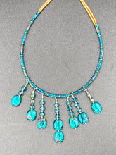 Load image into Gallery viewer, This necklace features vibrant teal quartzite and glass beads. The quartzite beads have a captivating swirl pattern, combining dark red and dark blue hues. It has an ethereal feel thanks to the various shades of blues, golds, and greens used to create a two-strand beaded design. For a dramatic finish, seven strands are added, completed with rectangular rainbow hematite beads for texture and depth. Both strands are held together with double-hole flat matte gold beads to keep a collar shape.