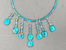 Load image into Gallery viewer, This necklace features vibrant teal quartzite and glass beads. The quartzite beads have a captivating swirl pattern, combining dark red and dark blue hues. It has an ethereal feel thanks to the various shades of blues, golds, and greens used to create a two-strand beaded design. For a dramatic finish, seven strands are added, completed with rectangular rainbow hematite beads for texture and depth. Both strands are held together with double-hole flat matte gold beads to keep a collar shape.