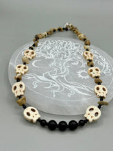 Load image into Gallery viewer, This skully necklace is full of neutral tones and is adorned with natural black obsidian and bamboo chips, as well as small glass beads. It has a unique design that creates a striking silhouette across the wearer's neck. An ideal statement piece for bold fashion enthusiasts. Measurements: Necklace - 19 1/4"