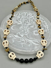 Load image into Gallery viewer, This skully necklace is full of neutral tones and is adorned with natural black obsidian and bamboo chips, as well as small glass beads. It has a unique design that creates a striking silhouette across the wearer's neck. An ideal statement piece for bold fashion enthusiasts. Measurements: Necklace - 19 1/4"