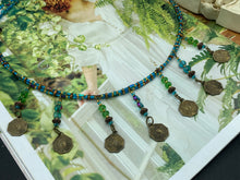 Load image into Gallery viewer, This necklace features vivid green and blue glass beading and a yin yang brass octagon charm. Each side of the charm is distinct, creating a two-in-one look. The look is further enhanced with the addition of rectangular rainbow hematite beads for added visual complexity. Double-hole flat matte gold beads keep the collar shape and add to the necklace's elegant draping when worn.  Measurements:    Necklace is 23” in length