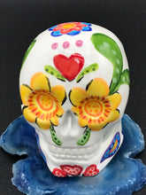 Load image into Gallery viewer, Inspired by nature, this ceramic skull statuette is full of vibrant color and intricate details. The primary color is white with multi-color floral motifs that are painted across the skull, along with hearts and leaves and lively colorful accents around the entire skull.