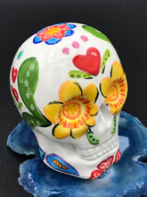 Load image into Gallery viewer, Inspired by nature, this ceramic skull statuette is full of vibrant color and intricate details. The primary color is white with multi-color floral motifs that are painted across the skull, along with hearts and leaves and lively colorful accents around the entire skull.