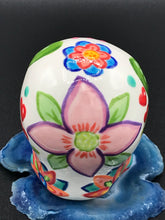 Load image into Gallery viewer, Inspired by nature, this ceramic skull statuette is full of vibrant color and intricate details. The primary color is white with multi-color floral motifs that are painted across the skull, along with hearts and leaves and lively colorful accents around the entire skull.