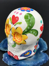 Load image into Gallery viewer, Inspired by nature, this ceramic skull statuette is full of vibrant color and intricate details. The primary color is white with multi-color floral motifs that are painted across the skull, along with hearts and leaves and lively colorful accents around the entire skull.