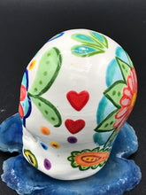 Load image into Gallery viewer, Inspired by nature, this ceramic skull statuette is full of vibrant color and intricate details. The primary color is white with multi-color floral motifs that are painted across the