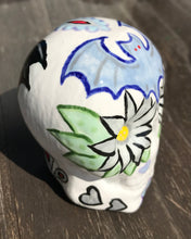 Load image into Gallery viewer, This ceramic skull art is a unique and eye-catching combination of bright colors and dark tones. It features two bat silhouettes and flowers in warm colors like red, yellow, and green, as well as medium gray and royal blue. Its vibrant red eyes give the piece an inviting touch. Perfect for a mini goth witchy altar, this figurine is sure to stand out. Measurements: H 2 3/4” x W 2” x D 2”