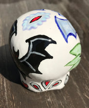 Load image into Gallery viewer, This ceramic skull art is a unique and eye-catching combination of bright colors and dark tones. It features two bat silhouettes and flowers in warm colors like red, yellow, and green, as well as medium gray and royal blue. Its vibrant red eyes give the piece an inviting touch. Perfect for a mini goth witchy altar, this figurine is sure to stand out. Measurements: H 2 3/4” x W 2” x D 2”