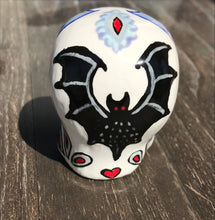 Load image into Gallery viewer, This ceramic skull art is a unique and eye-catching combination of bright colors and dark tones. It features two bat silhouettes and flowers in warm colors like red, yellow, and green, as well as medium gray and royal blue. Its vibrant red eyes give the piece an inviting touch. Perfect for a mini goth witchy altar, this figurine is sure to stand out. Measurements: H 2 3/4” x W 2” x D 2”
