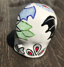Load image into Gallery viewer, This ceramic skull art is a unique and eye-catching combination of bright colors and dark tones. It features two bat silhouettes and flowers in warm colors like red, yellow, and green, as well as medium gray and royal blue. Its vibrant red eyes give the piece an inviting touch. Perfect for a mini goth witchy altar, this figurine is sure to stand out. Measurements: H 2 3/4” x W 2” x D 2”