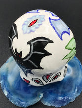 Load image into Gallery viewer, This ceramic skull art is a unique and eye-catching combination of bright colors and dark tones. It features two bat silhouettes and flowers in warm colors like red, yellow, and green, as well as medium gray and royal blue. Its vibrant red eyes give the piece an inviting touch. Perfect for a mini goth witchy altar, this figurine is sure to stand out. Measurements: H 2 3/4” x W 2” x D 2”