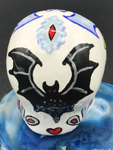 Load image into Gallery viewer, This ceramic skull art is a unique and eye-catching combination of bright colors and dark tones. It features two bat silhouettes and flowers in warm colors like red, yellow, and green, as well as medium gray and royal blue. Its vibrant red eyes give the piece an inviting touch. Perfect for a mini goth witchy altar, this figurine is sure to stand out. Measurements: H 2 3/4” x W 2” x D 2”