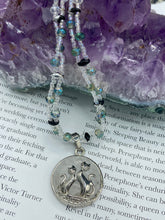 Load image into Gallery viewer, This stylish necklace features a shimmery sterling silver quartz pendant with cats and stars, making it the perfect accessory for any cat lover. With two-hole silver and black beads that stand out against the light aquamarine glass beads, this double strand necklace holds a round kitty pendant made with various hues of clear and iridescent glass beads. Add a touch of purrfection to your look with this statement piece. Measurements: Necklace is 15”adjustable up to 18"