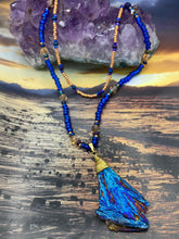 Load image into Gallery viewer, A bold deep blue kyanite wired stone pendant is surrounded by two strands of glass bead necklaces featuring a deep blue and gold shimmer palette. The shorter strand is designed to be worn as a collar or choker, while the longer second strand creates a striking contrast. With its bold appearance and eye-catching gemstone pendant, this necklace is a must-have for any jewelry collection. Necklace is 15” in length, can be adjusted to 18"