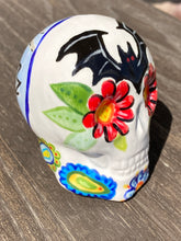 Load image into Gallery viewer, This Bat Lovers Ceramic Skull Art is a unique addition to any dark gothic collection. Featuring a mini batty with bright red eyes and outlined in royal blue, accented with white lines to highlight its small bones and an additional bat with vibrant red eyes on the opposite side. Enjoy the playful contrast of warm tones and a darker theme in this eye-catching figurine. Measurements: H 2 3/4” x W 2” x D 2”