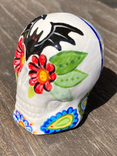 Load image into Gallery viewer, This Bat Lovers Ceramic Skull Art is a unique addition to any dark gothic collection. Featuring a mini batty with bright red eyes and outlined in royal blue, accented with white lines to highlight its small bones and an additional bat with vibrant red eyes on the opposite side. Enjoy the playful contrast of warm tones and a darker theme in this eye-catching figurine. Measurements: H 2 3/4” x W 2” x D 2”