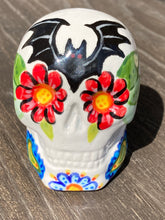 Load image into Gallery viewer, This Bat Lovers Ceramic Skull Art is a unique addition to any dark gothic collection. Featuring a mini batty with bright red eyes and outlined in royal blue, accented with white lines to highlight its small bones and an additional bat with vibrant red eyes on the opposite side. Enjoy the playful contrast of warm tones and a darker theme in this eye-catching figurine. Measurements: H 2 3/4” x W 2” x D 2”