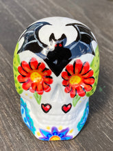 Load image into Gallery viewer, Inspired by nature, this ceramic skull statuette is full of vibrant color and intricate details. The primary color is white with a batty for bats theme and with multi-color floral motifs, along with hearts and leaves and lively colorful accents around the entire skull. Measurements: H 2 3/4” x W 2” x D 2”