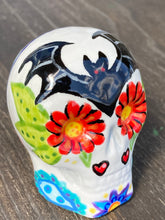 Load image into Gallery viewer, Inspired by nature, this ceramic skull statuette is full of vibrant color and intricate details. The primary color is white with a batty for bats theme and with multi-color floral motifs, along with hearts and leaves and lively colorful accents around the entire skull. Measurements: H 2 3/4” x W 2” x D 2”