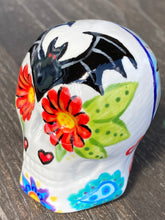 Load image into Gallery viewer, Inspired by nature, this ceramic skull statuette is full of vibrant color and intricate details. The primary color is white with a batty for bats theme and with multi-color floral motifs, along with hearts and leaves and lively colorful accents around the entire skull. Measurements: H 2 3/4” x W 2” x D 2”
