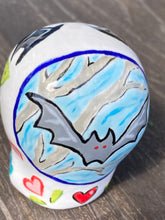 Load image into Gallery viewer, Inspired by nature, this ceramic skull statuette is full of vibrant color and intricate details. The primary color is white with a batty for bats theme and with multi-color floral motifs, along with hearts and leaves and lively colorful accents around the entire skull. Measurements: H 2 3/4” x W 2” x D 2”