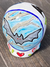 Load image into Gallery viewer, Inspired by nature, this ceramic skull statuette is full of vibrant color and intricate details. The primary color is white with a batty for bats theme and with multi-color floral motifs, along with hearts and leaves and lively colorful accents around the entire skull. Measurements: H 2 3/4” x W 2” x D 2”
