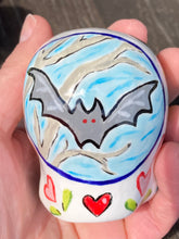 Load image into Gallery viewer, Inspired by nature, this ceramic skull statuette is full of vibrant color and intricate details. The primary color is white with a batty for bats theme and with multi-color floral motifs, along with hearts and leaves and lively colorful accents around the entire skull. Measurements: H 2 3/4” x W 2” x D 2”