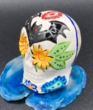 Load image into Gallery viewer, Inspired by nature, this ceramic skull statuette is full of vibrant color and intricate details. The primary color is white with a batty for bats theme and with multi-color floral motifs, along with hearts and leaves and lively colorful accents around the entire skull. Measurements: H 2 3/4” x W 2” x D 2”