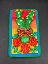 Load image into Gallery viewer, This handcrafted polymer clay wall art features a wood frame painted in a gradient from light yellow to dark red, creating a stunning sunset look. With a bright green skull cactus plant and bright yellow blooming flowers. Boasting vivid marigold blooms and a bright teal and green hue frame to make a bold contrast with the warm background hue. this piece is sure to make a statement. Measurements: