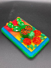 Load image into Gallery viewer, This handcrafted polymer clay wall art features a wood frame painted in a gradient from light yellow to dark red, creating a stunning sunset look. With a bright green skull cactus plant and bright yellow blooming flowers. Boasting vivid marigold blooms and a bright teal and green hue frame to make a bold contrast with the warm background hue. this piece is sure to make a statement. Measurements:
