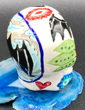 Load image into Gallery viewer, Inspired by nature, this ceramic skull statuette is full of vibrant color and intricate details. The primary color is white with a batty for bats theme and with multi-color floral motifs, along with hearts and leaves and lively colorful accents around the entire skull. Measurements: H 2 3/4” x W 2” x D 2”