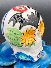 Load image into Gallery viewer, Inspired by nature, this ceramic skull statuette is full of vibrant color and intricate details. The primary color is white with a batty for bats theme and with multi-color floral motifs, along with hearts and leaves and lively colorful accents around the entire skull. Measurements: H 2 3/4” x W 2” x D 2”