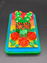 Load image into Gallery viewer, This handcrafted polymer clay wall art features a wood frame painted in a gradient from light yellow to dark red, creating a stunning sunset look. With a bright green skull cactus plant and bright yellow blooming flowers. Boasting vivid marigold blooms and a bright teal and green hue frame to make a bold contrast with the warm background hue. this piece is sure to make a statement. Measurements: